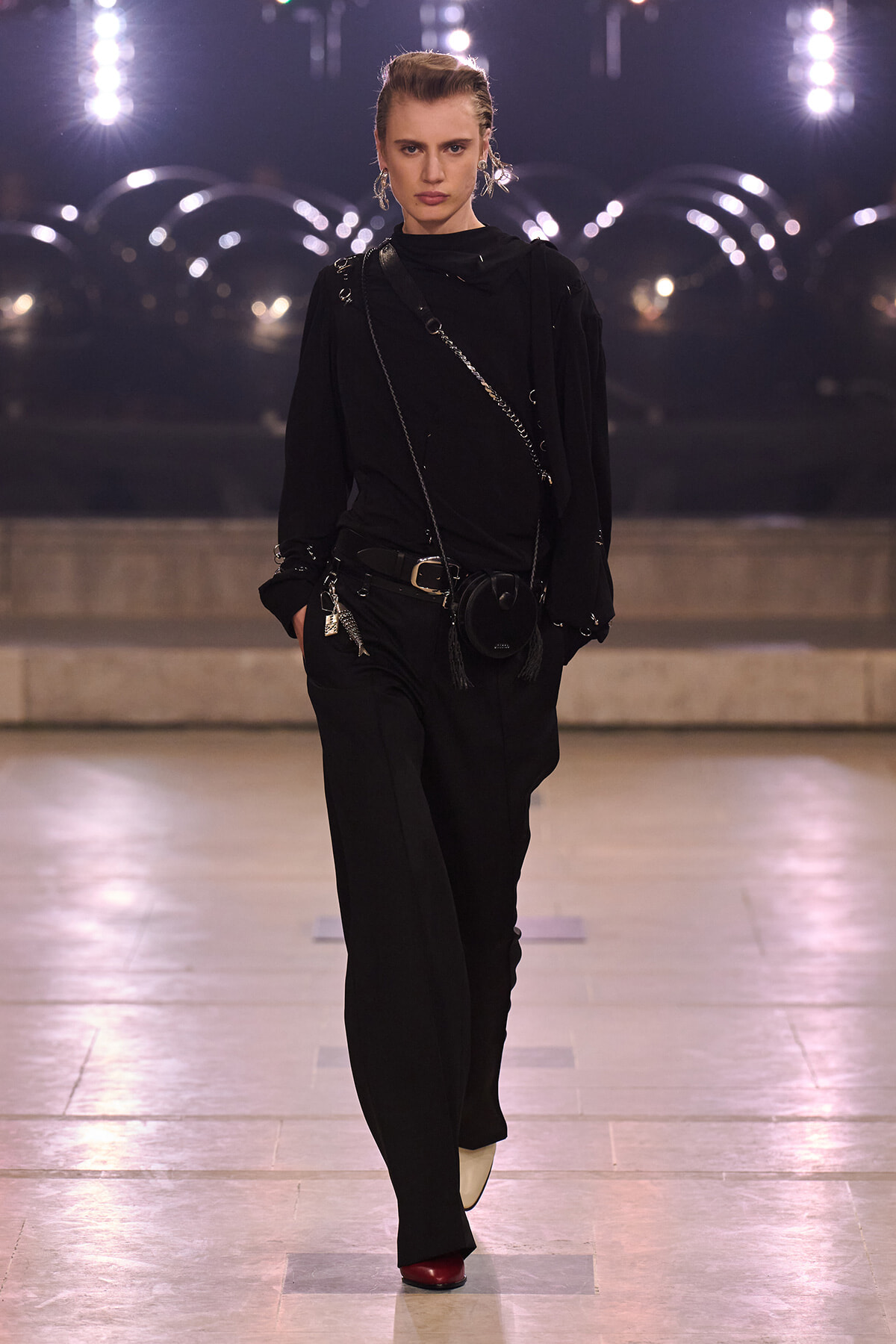 Model walking a runway in a black outfit with a crossbody chain and small black purse, spotlighted on stage.