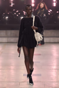 Model walking a runway in a black knit mini dress with a waist tie, carrying a cream crossbody bag and wearing studded sock-boot heels.
