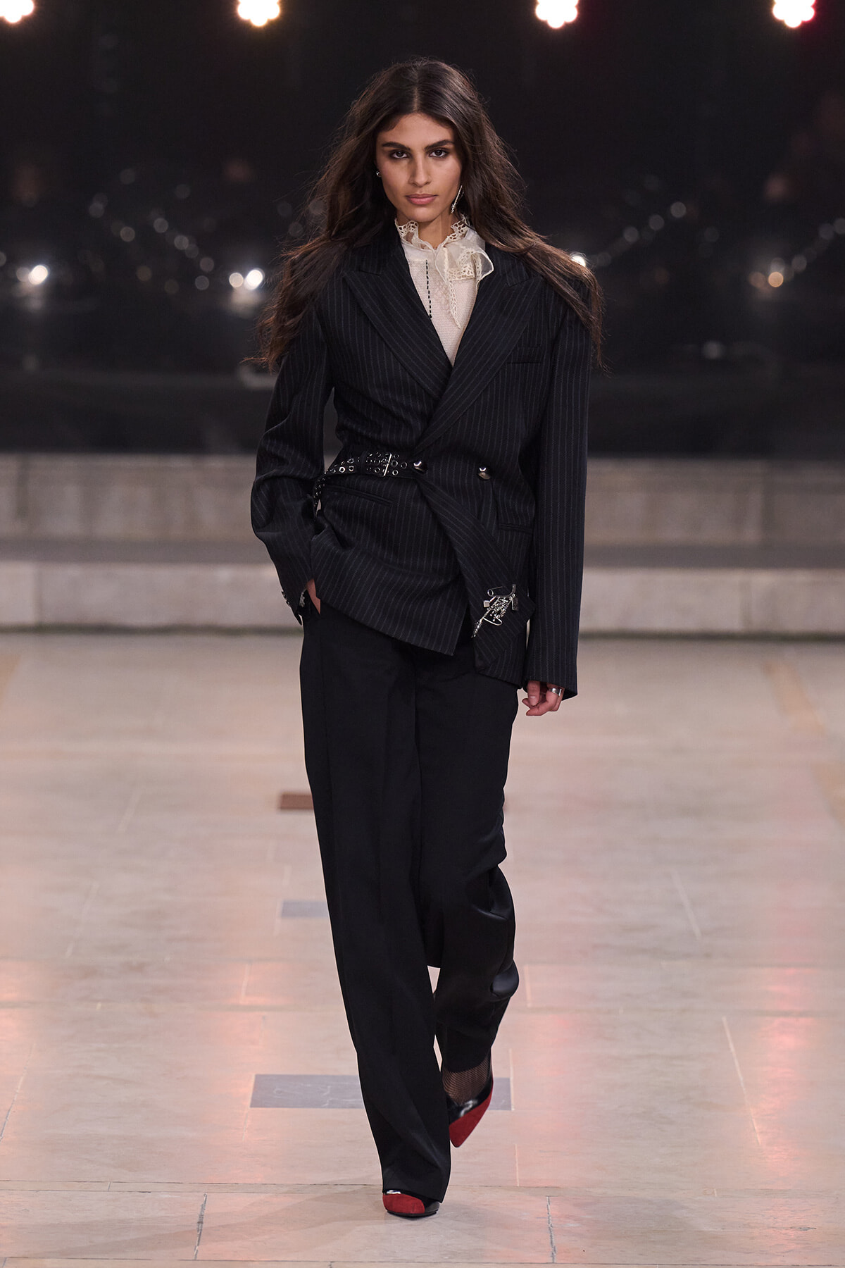 Model on a runway wearing a belted pinstripe blazer, lace high-neck blouse, and wide-leg black trousers with red shoes.
