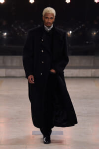 Male model on a runway wearing a long black double-breasted wool coat, black outfit, and black boots.