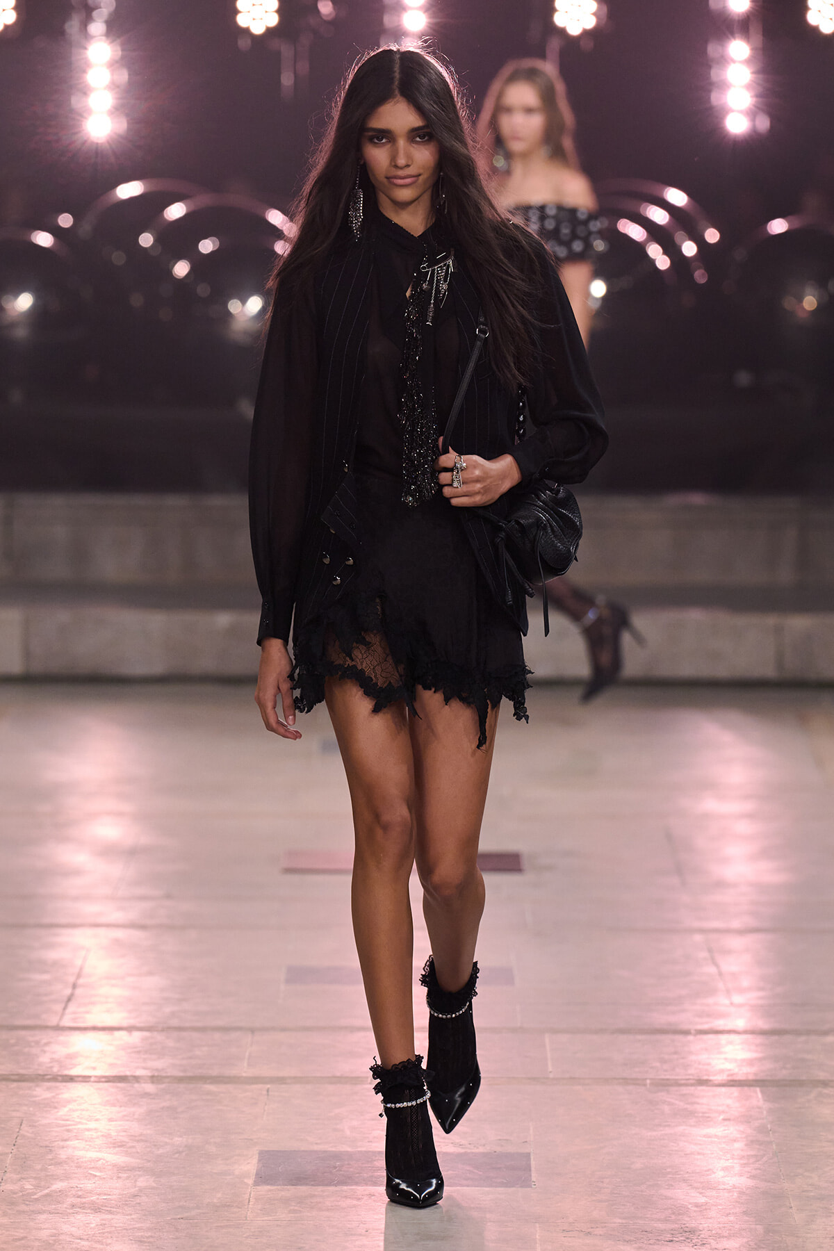Model walking a runway in an all-black ensemble with a lace-trimmed mini skirt, sheer blouse, and pinstripe vest, carrying a black bag under bright stage lights.