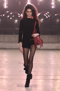 Model walking down a fashion show runway in a black knit top, black shorts, dotted sheer tights, and studded boots, carrying a red bag with fringe.