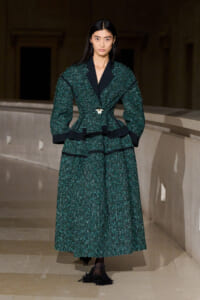 Female model on a runway wearing a dark green textured suit with black lapels and a gold brooch at the waist.