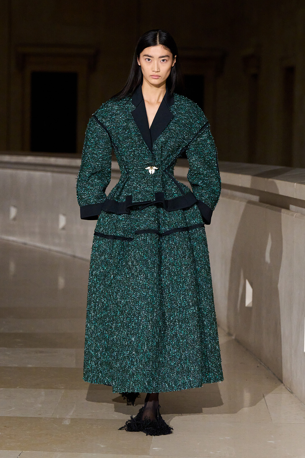 Female model on a runway wearing a dark green textured suit with black lapels and a gold brooch at the waist.