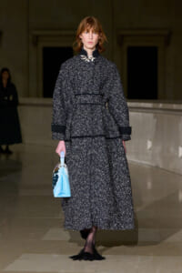 Model on a runway wearing a textured black-and-white speckled coat and skirt with black trim, carrying a light blue handbag.
