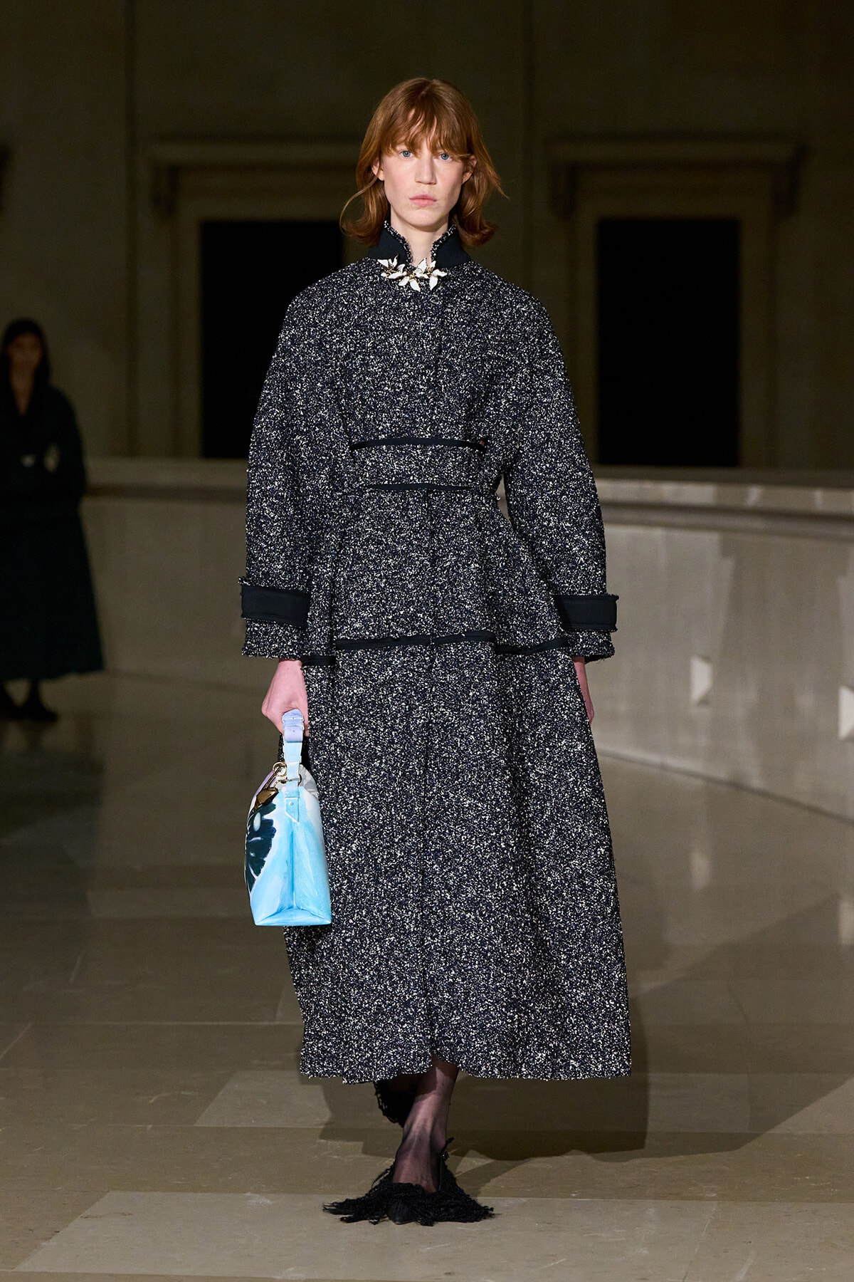 Model on a runway wearing a textured black-and-white speckled coat and skirt with black trim, carrying a light blue handbag.