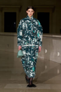 Model on a runway wearing a teal distressed tweed suit with fringe, carrying a mint green handbag.