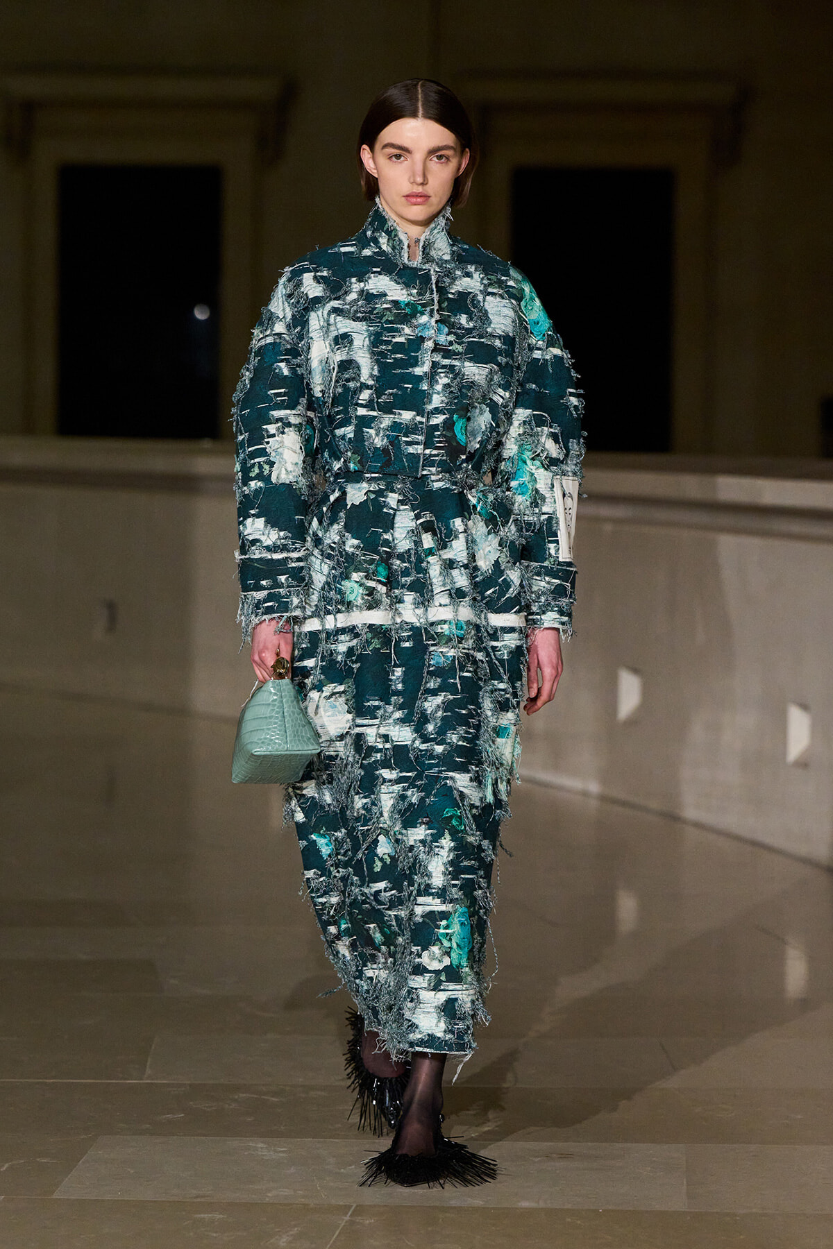 Model on a runway wearing a teal distressed tweed suit with fringe, carrying a mint green handbag.
