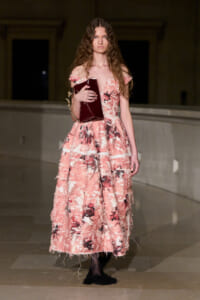 Model wearing a pink floral off‑shoulder gown on a runway, holding a burgundy clutch.