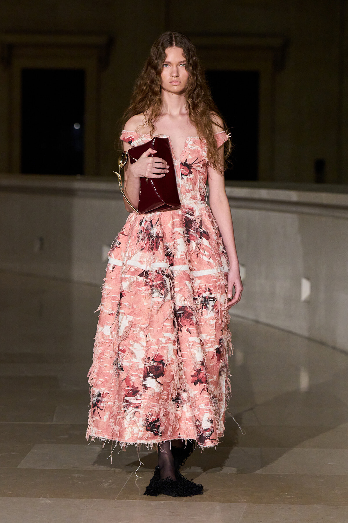 Model wearing a pink floral off‑shoulder gown on a runway, holding a burgundy clutch.