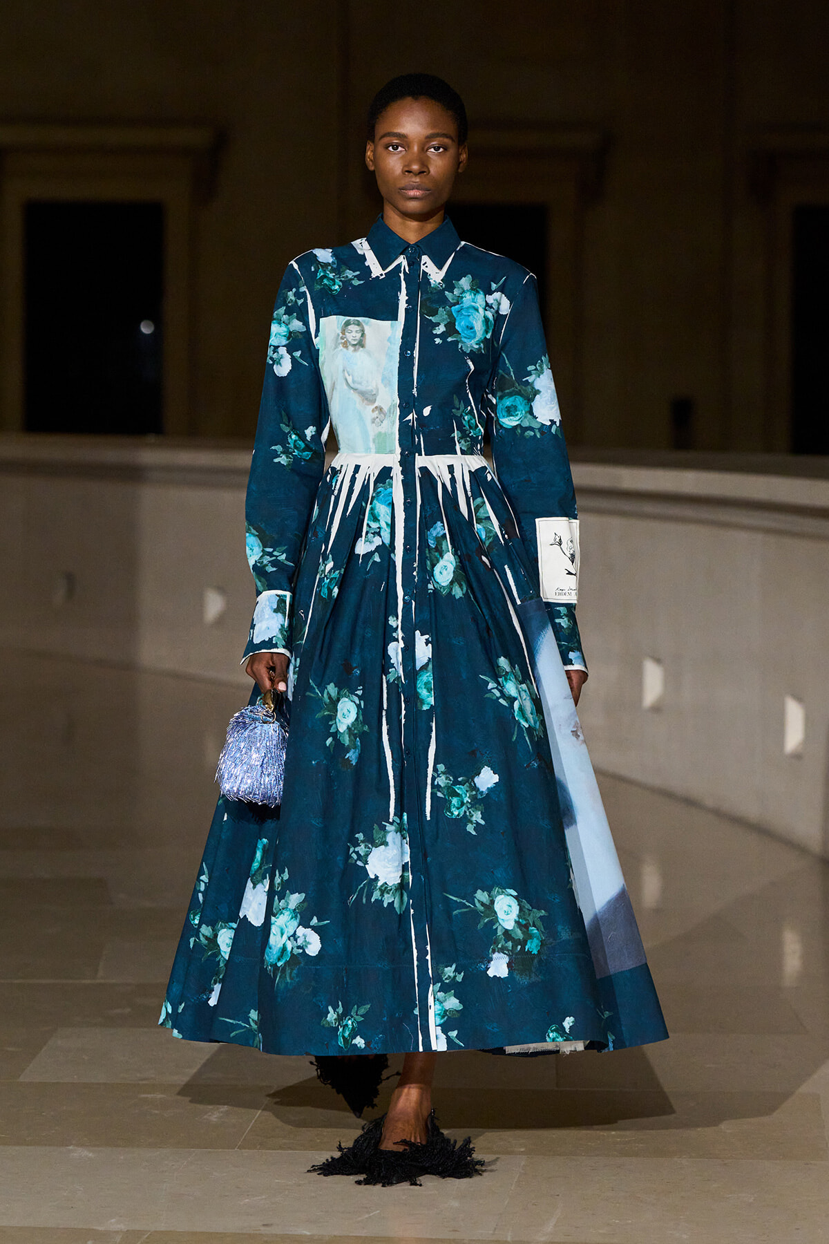 Model walking a runway in a long teal floral print dress with white stripe accents and a small blue fringed handbag.