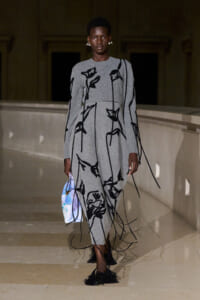 Model on a runway wearing a gray knit dress with black abstract floral lines and long fringe tassels, carrying a pastel blue and white purse.