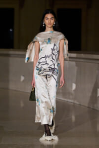 Model walks a runway in a silky, off-white floral gown with cape sleeves and fringe hem, carrying a small green handbag.