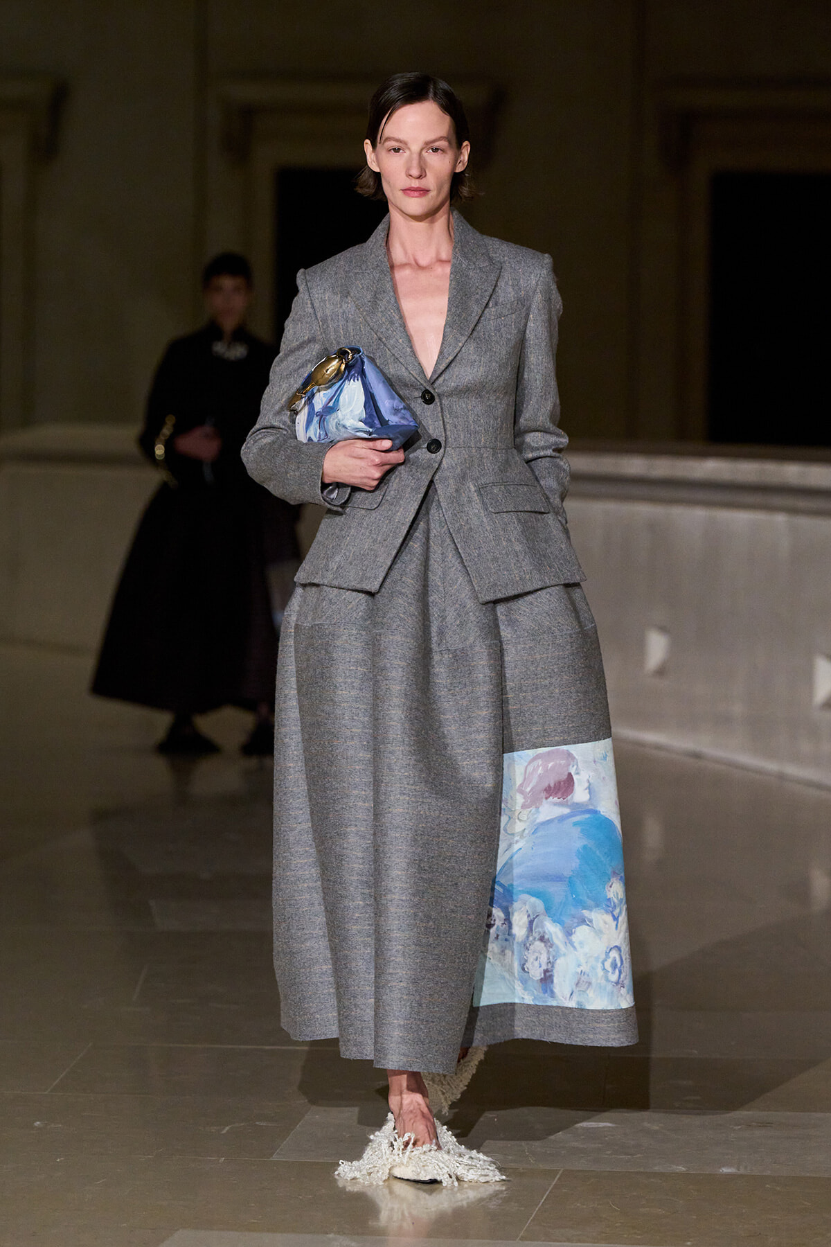 Model walks a fashion show in a tailored gray suit with a pleated, layered skirt; she carries a blue satin clutch and wears fringed white shoes.