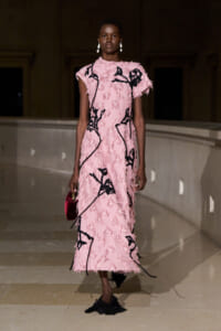 Model walking a runway in a pink textured fringe dress with black embroidery, carrying a small red handbag and wearing white drop earrings.