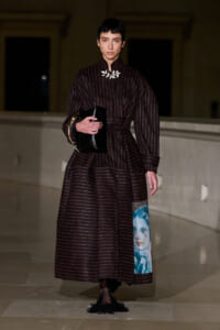 Female model in a dark burgundy striped, full-length coat-like dress, belt cinched, holding a glossy black clutch; white floral necklace and blue portrait panel on the skirt show fashion details.