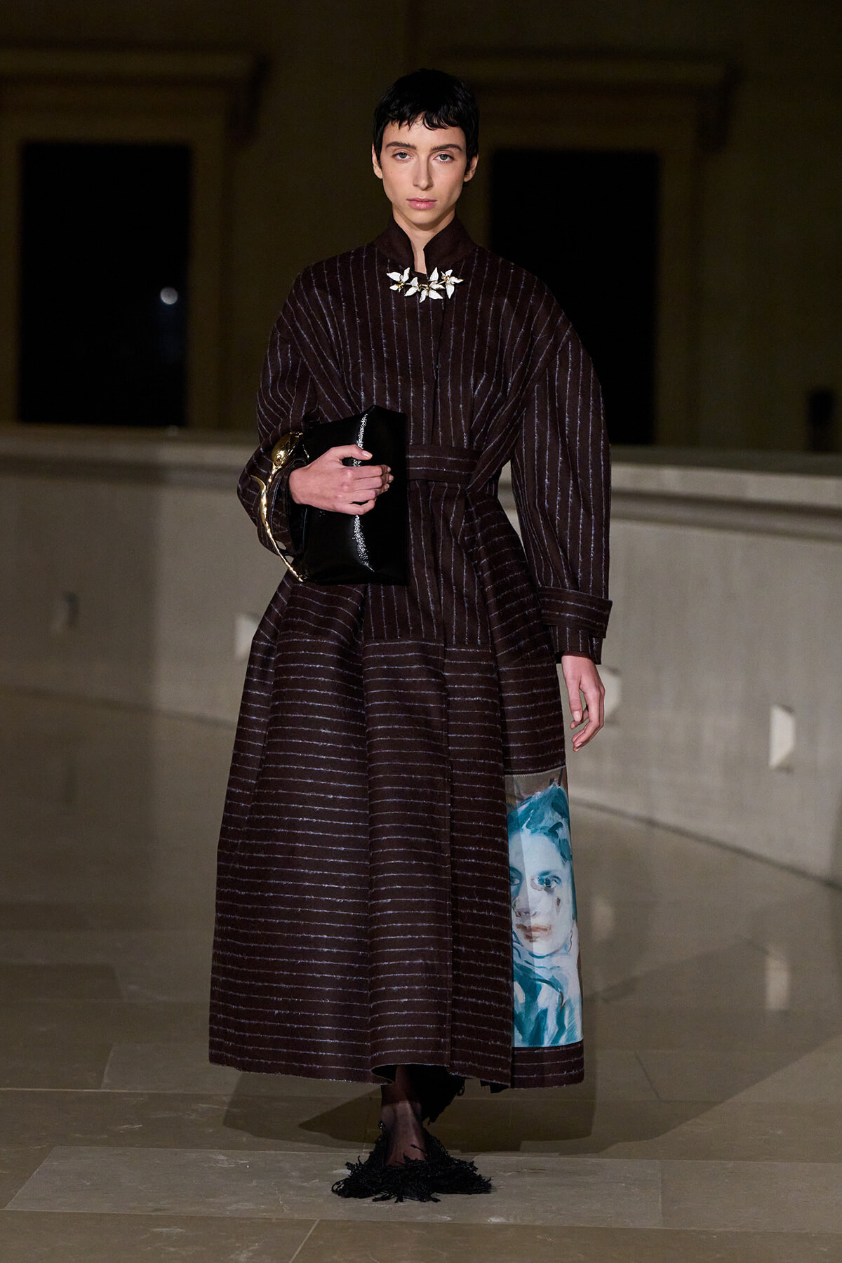 Female model in a dark burgundy striped, full-length coat-like dress, belt cinched, holding a glossy black clutch; white floral necklace and blue portrait panel on the skirt show fashion details.