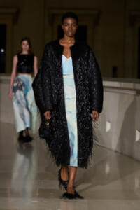 Model on a runway wearing a long black fringed coat over a pale blue dress; another model visible in the background at a fashion show.