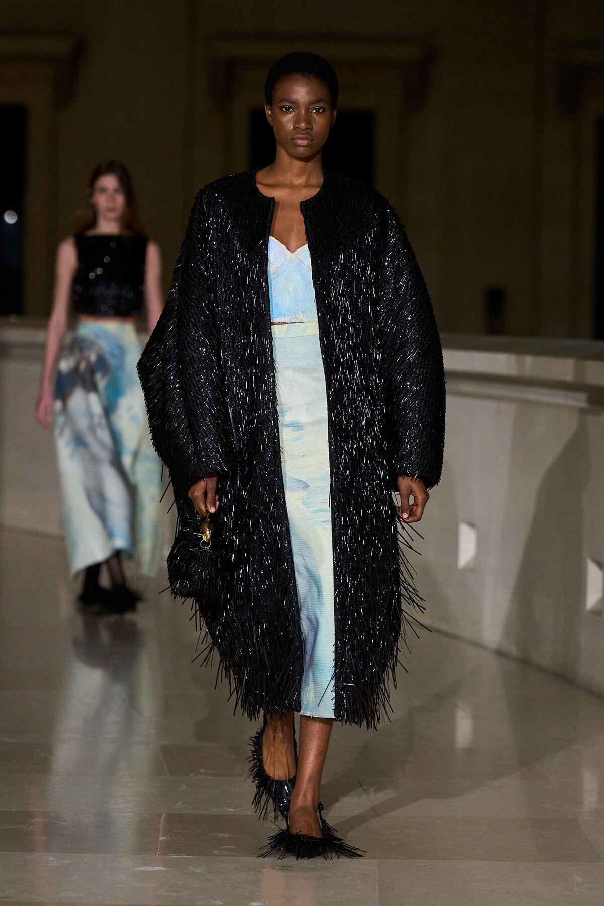 Model on a runway wearing a long black fringed coat over a pale blue dress; another model visible in the background at a fashion show.