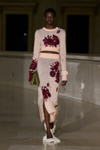 Model walking a runway in a pale pink knit dress with large burgundy floral appliqués and a belt at the waist, carrying a small green handbag.