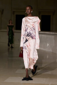 Model walking the runway in a pale pink, floral-print, oversized dress with fringe hems and a draped one-shoulder overlay, holding a small red handbag.