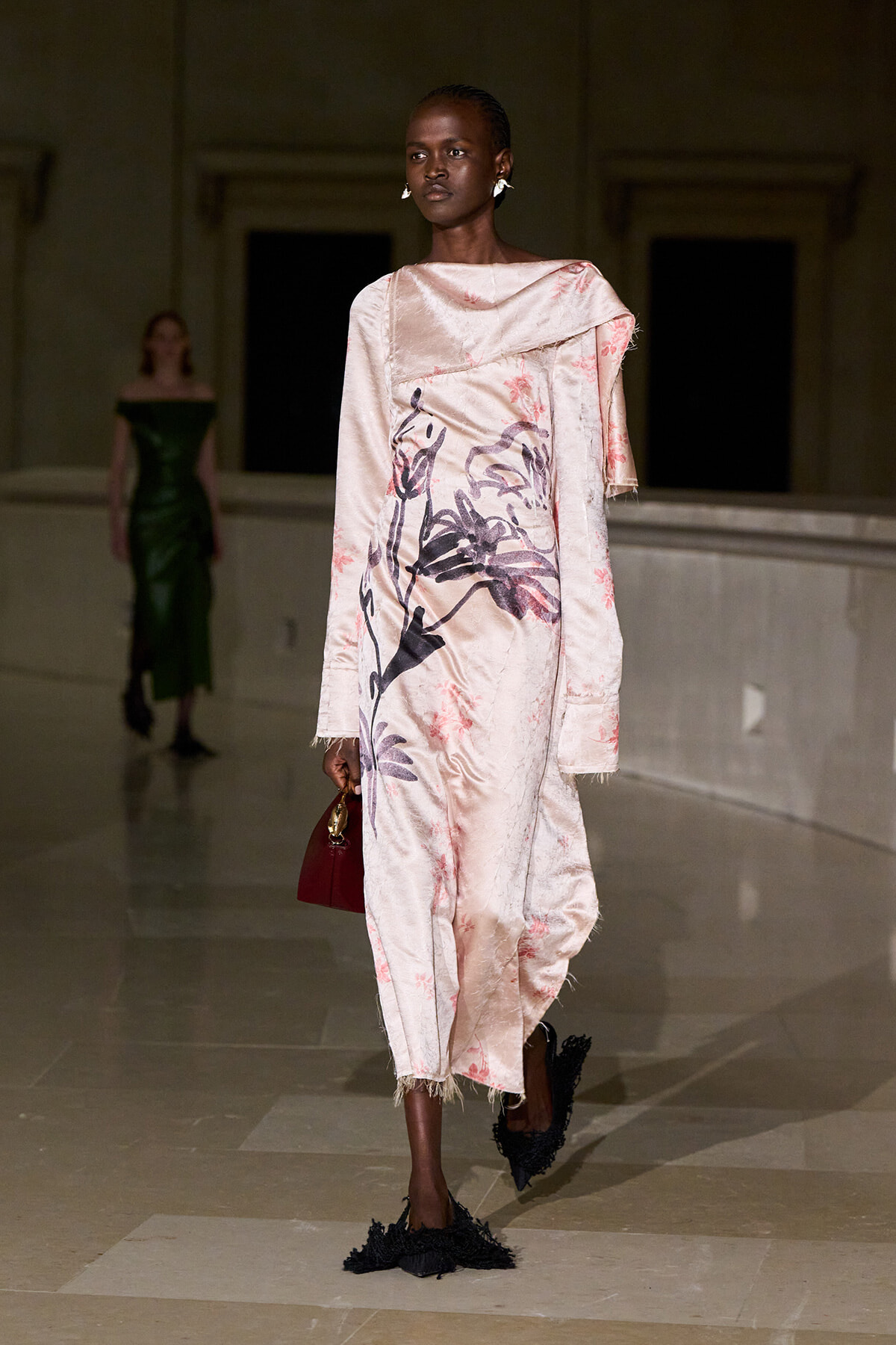 Model walking the runway in a pale pink, floral-print, oversized dress with fringe hems and a draped one-shoulder overlay, holding a small red handbag.