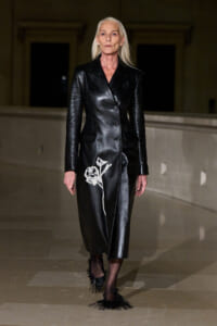 Model wears a black double-breasted leather coat with a white rose embroidery on the lower front, walking the runway.