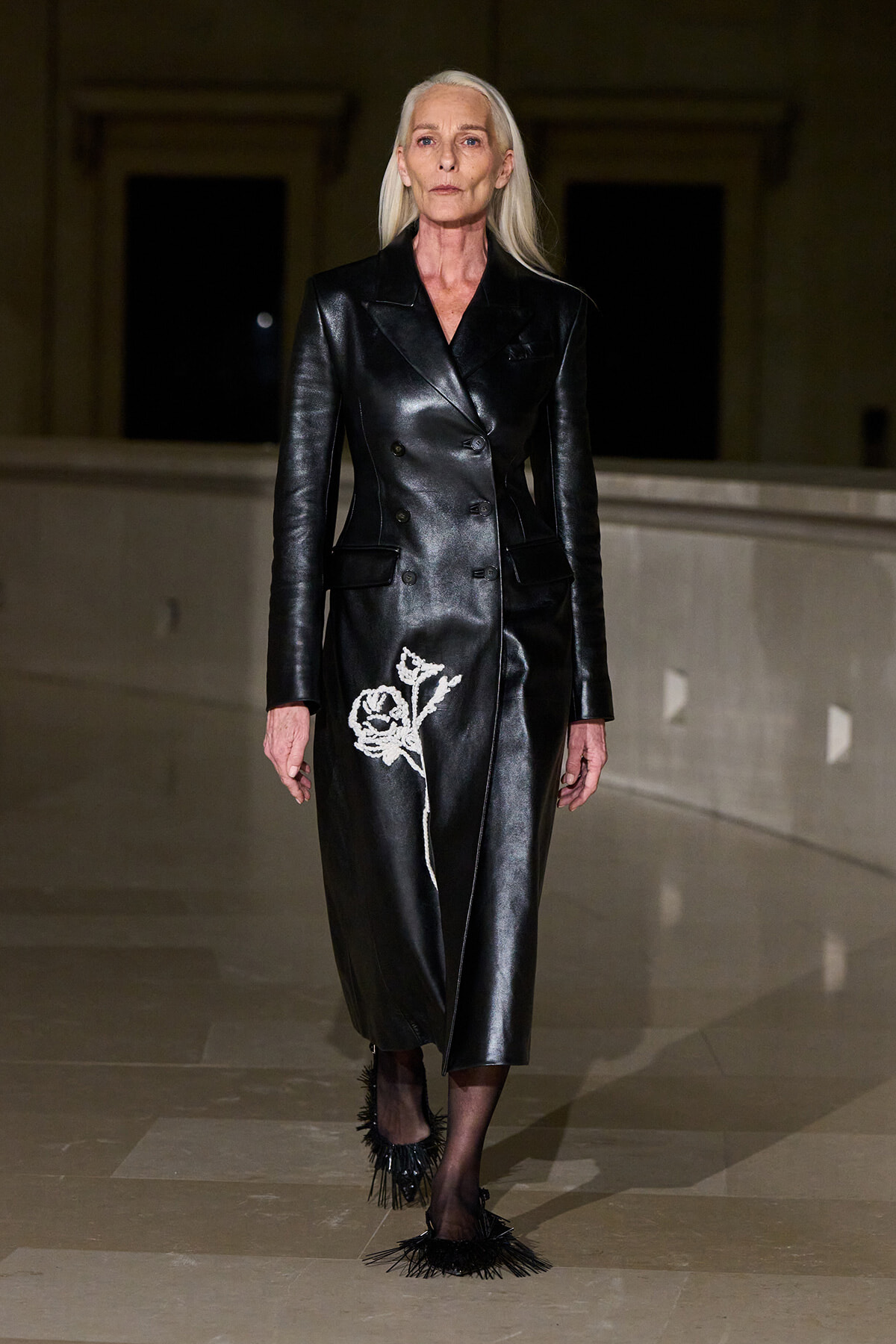 Model wears a black double-breasted leather coat with a white rose embroidery on the lower front, walking the runway.