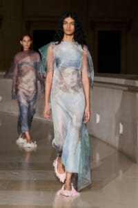 Model walking a runway in a sheer pastel gown with face-print artwork and pink fluffy footwear.