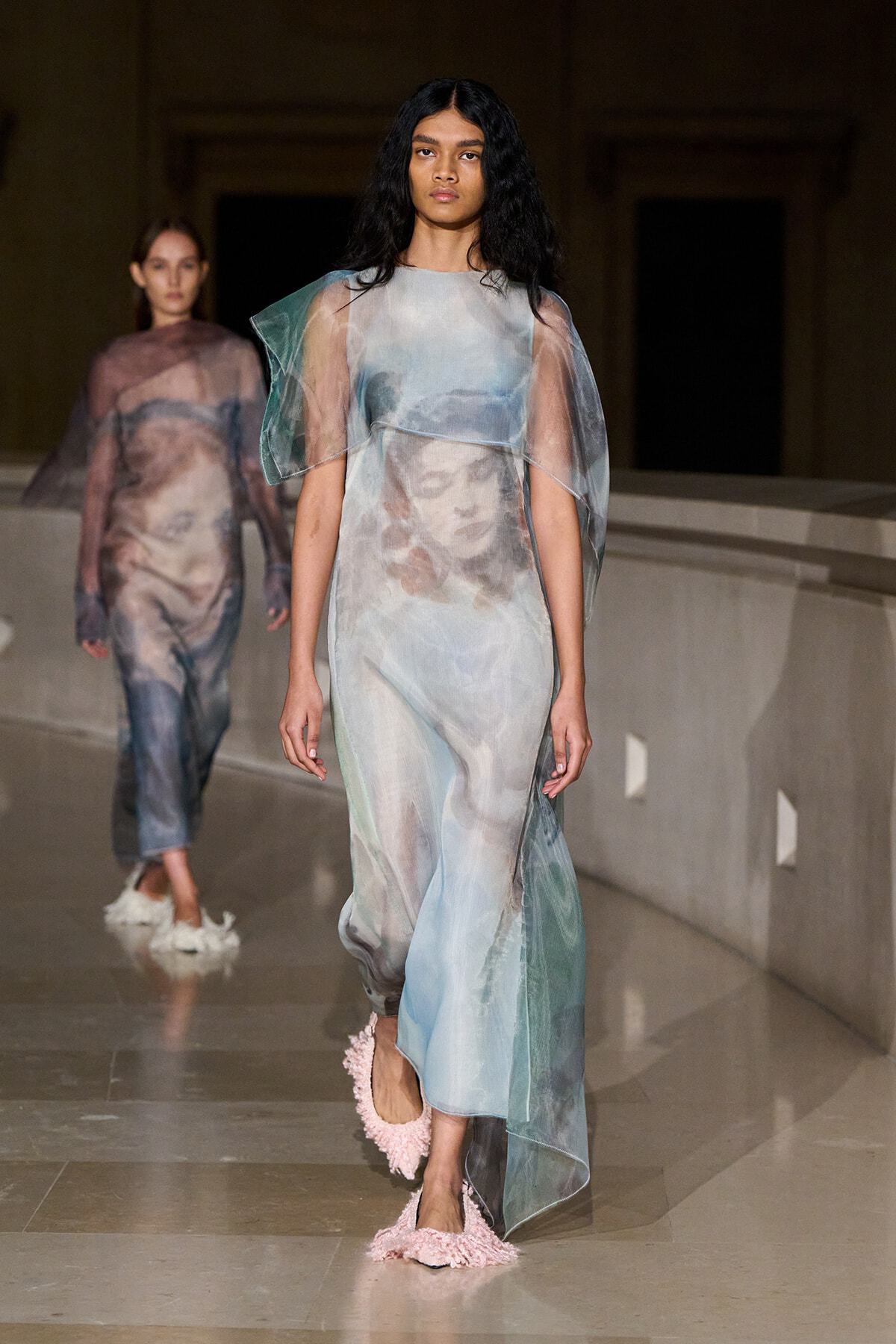 Model walking a runway in a sheer pastel gown with face-print artwork and pink fluffy footwear.