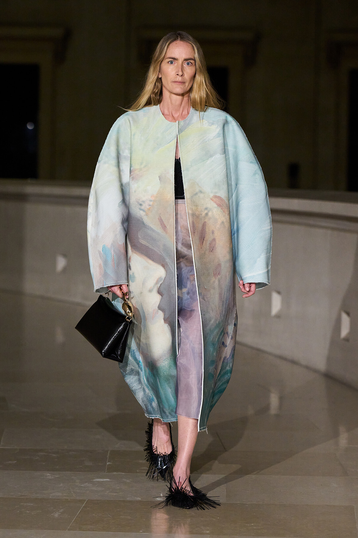 Model walking a runway in a loose pastel abstract coat over a black top and lavender skirt, carrying a small black handbag.