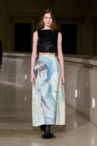 Model walking a runway in a black fringed crop top and a pastel watercolor-print maxi skirt on a dimly lit catwalk onlookers in the distance not visible.