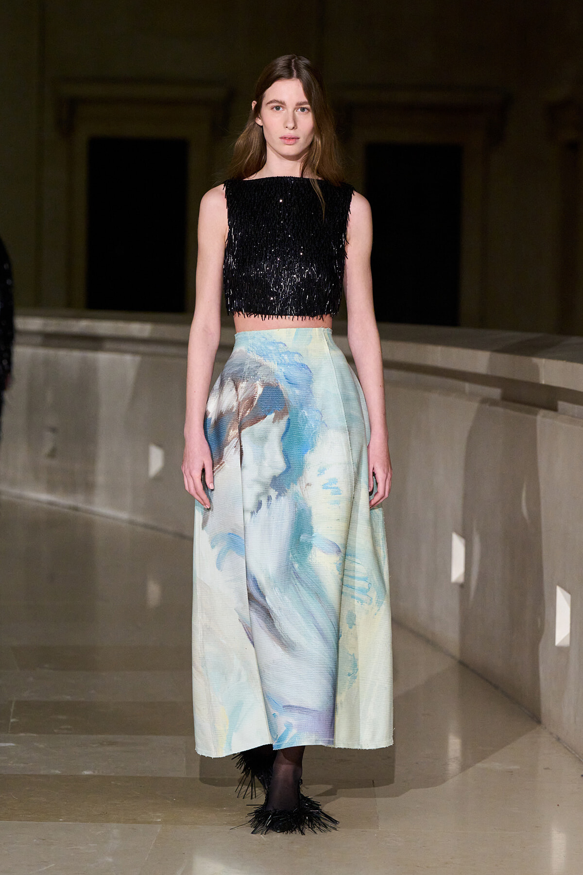 Model walking a runway in a black fringed crop top and a pastel watercolor-print maxi skirt on a dimly lit catwalk onlookers in the distance not visible.