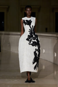 Model walks in a white sleeveless midi dress with bold black appliqué floral design down the side.