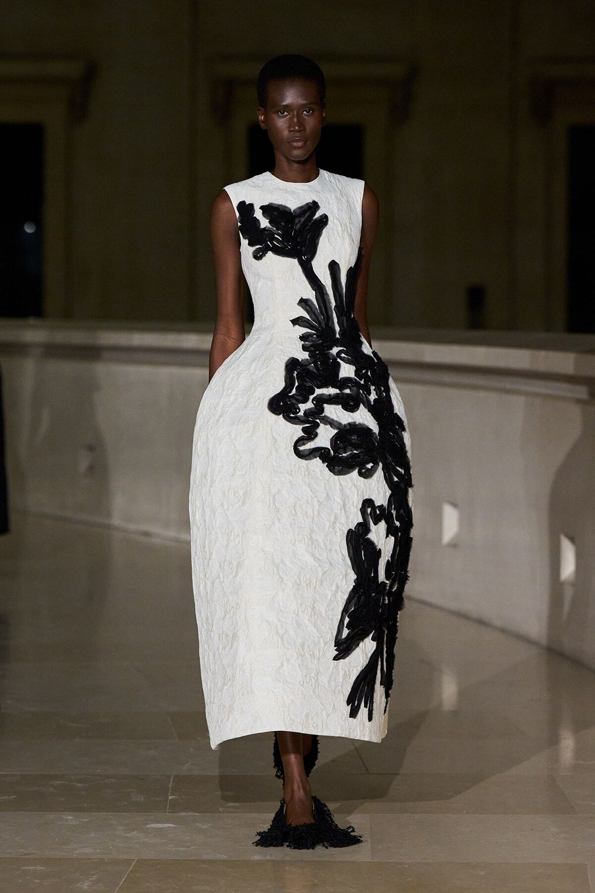 Model walks in a white sleeveless midi dress with bold black appliqué floral design down the side.
