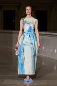 Model wearing a long sleeveless dress with blue abstract print walking on a runway, holding a small pink bag with blue fringe shoes visible at the bottom
