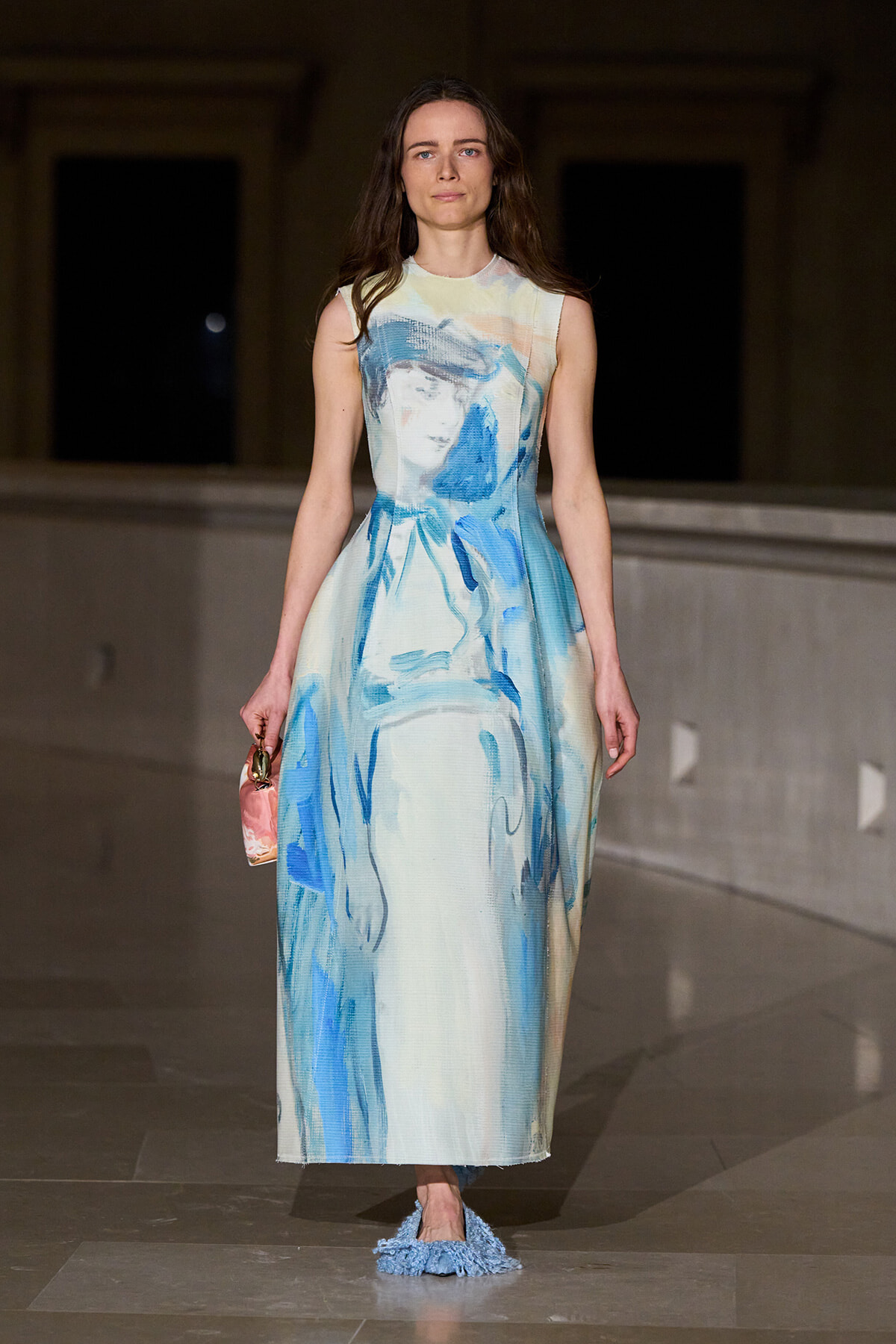 Model wearing a long sleeveless dress with blue abstract print walking on a runway, holding a small pink bag with blue fringe shoes visible at the bottom