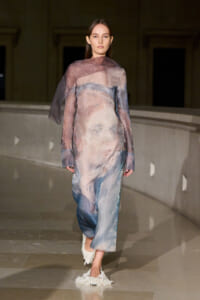 Model walking a fashion runway in a sheer pastel watercolor dress with blue, pink, and beige tones; translucent fabric and textured white shoes visible.