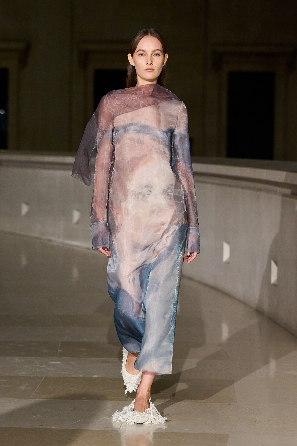 Model walking a fashion runway in a sheer pastel watercolor dress with blue, pink, and beige tones; translucent fabric and textured white shoes visible.