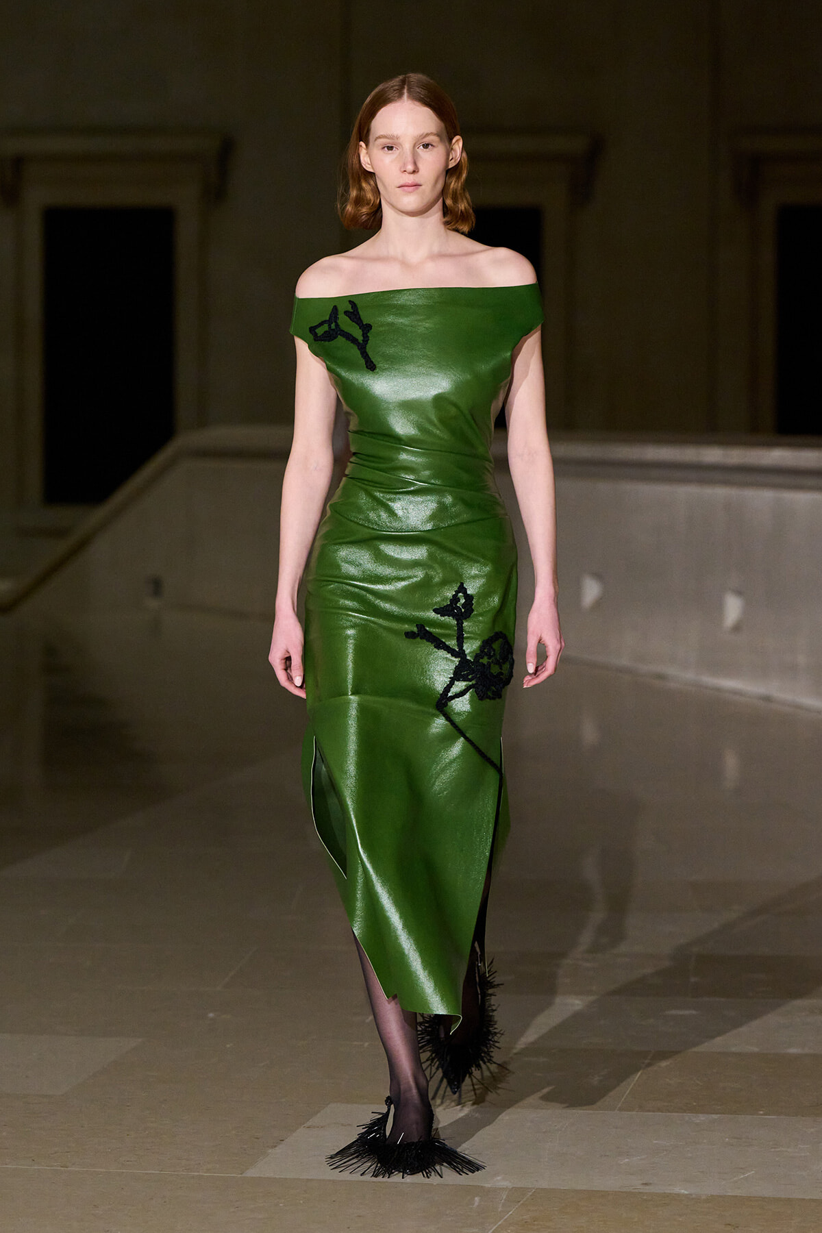 Model walking the runway in a strapless off‑the‑shoulder, emerald green leather midi dress with black embroidered motifs; black fringe heels and a neutral backstage setting.