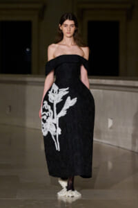 Model wearing an off-shoulder black lace gown with a large white floral applique on the left side, walking the runway.