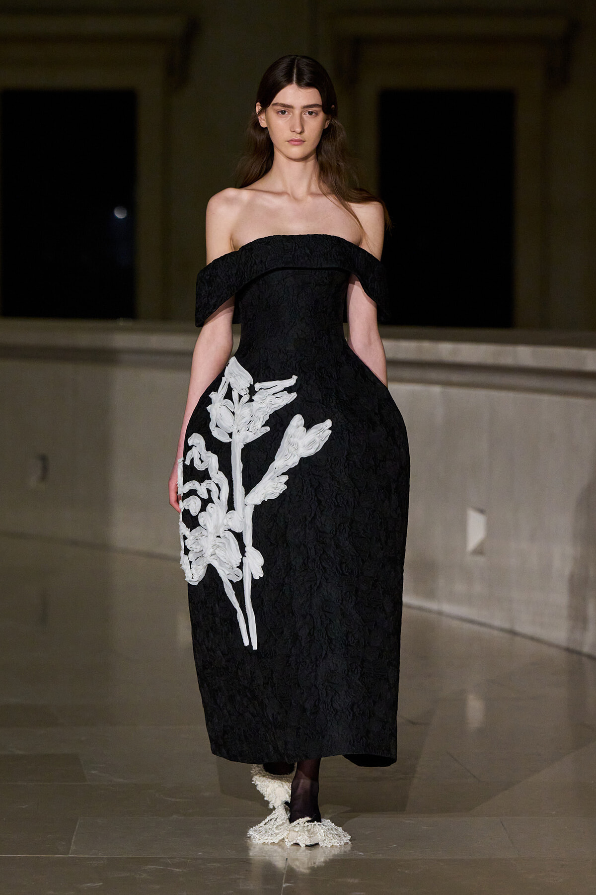 Model wearing an off-shoulder black lace gown with a large white floral applique on the left side, walking the runway.