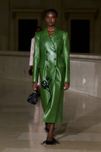 Model on a runway wearing a green double-breasted leather coat with black floral embroidery, carrying a small black handbag.