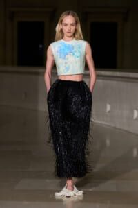 Model walks a runway in a pastel blue floral crop top and a long black fringed skirt. Beside her are white crocheted shoes visible at the floor level.