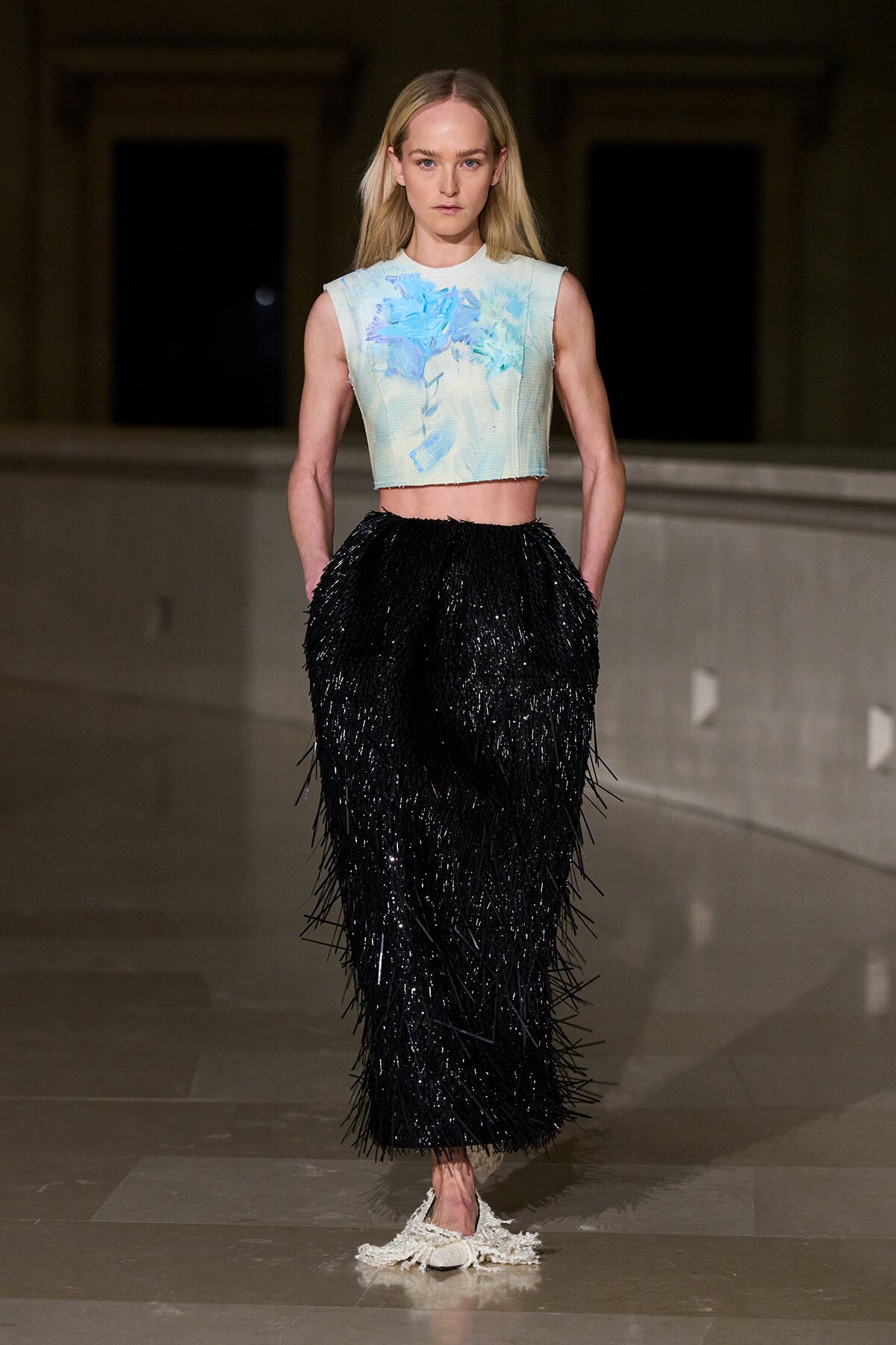 Model walks a runway in a pastel blue floral crop top and a long black fringed skirt. Beside her are white crocheted shoes visible at the floor level.