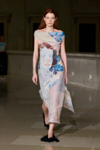 Model walking a runway in a pastel off-the-shoulder dress with an abstract print and flowing skirt.