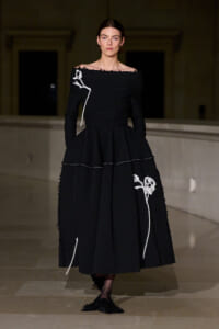 Model on a runway wearing a black off-shoulder textured gown with white embroidered floral lines and pockets.