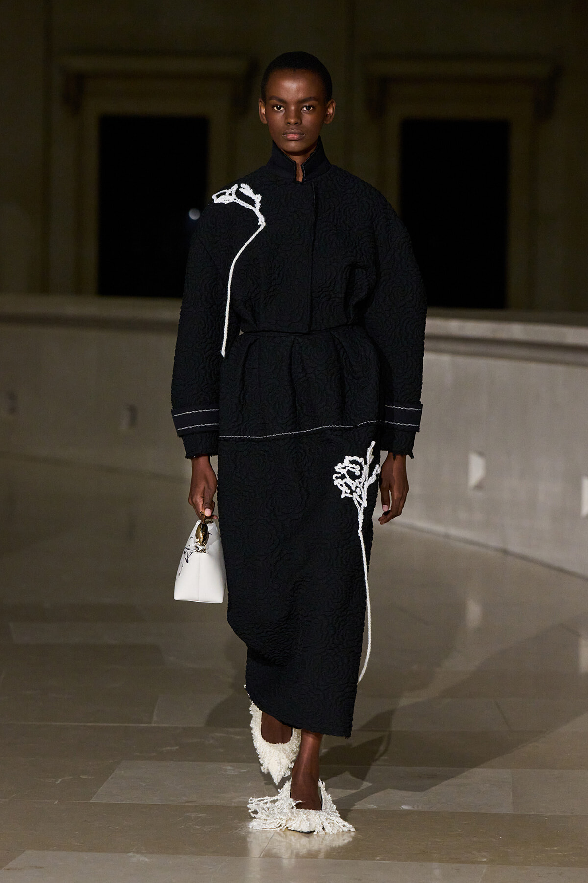 Model walking a runway in a black quilted outfit with white embroidered accents, carrying a small white handbag.