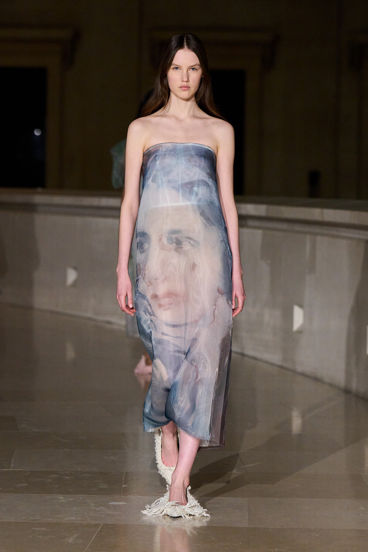 Strapless, floor-length dress with a large, faded portrait print on the fabric, worn by a model walking a runway at night.