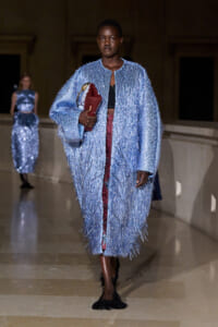 Model on a runway wearing a light-blue fringed coat, black top, burgundy pants, and black fringed shoes, clutching a red purse and a patterned bag.